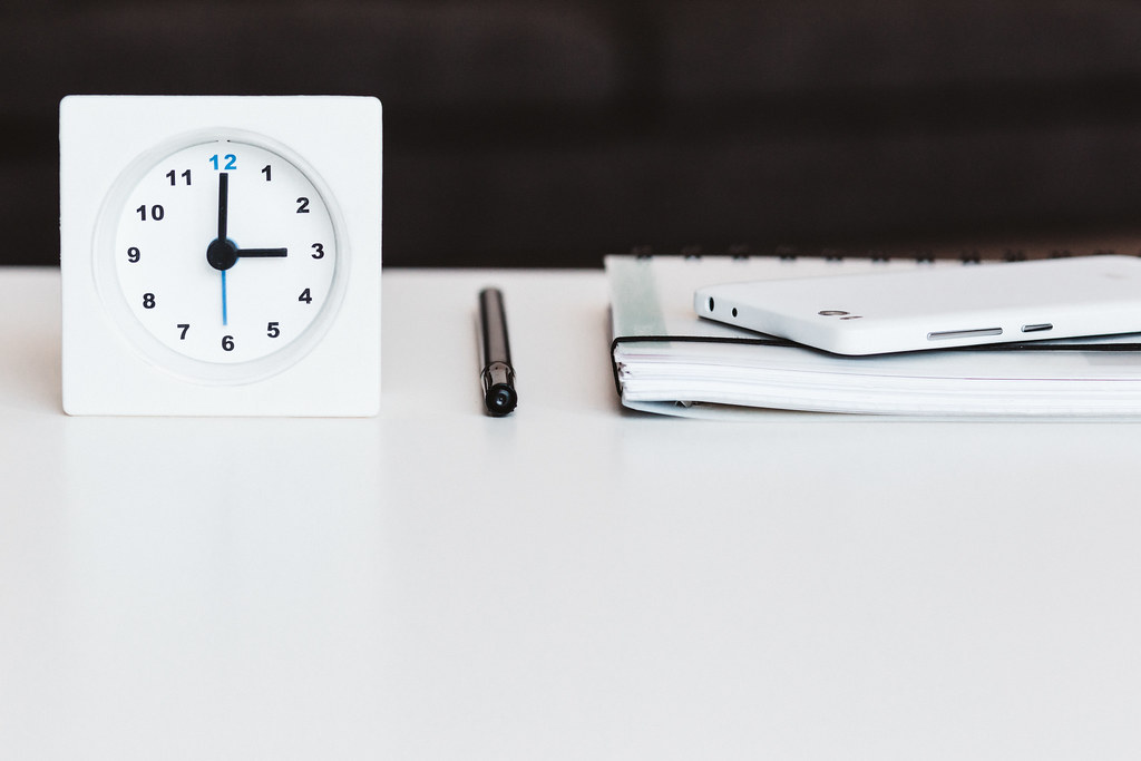 Mesa de oficina minimalista con un reloj analógico, un bolígrafo y un cuaderno con un smartphone encima, representando orden y planificación.