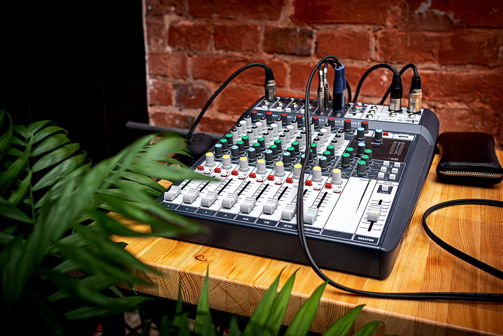 Una consola de mezclas de audio profesional situada sobre una mesa de madera con cables conectados, con una planta verde en primer plano y una pared de ladrillo al fondo.