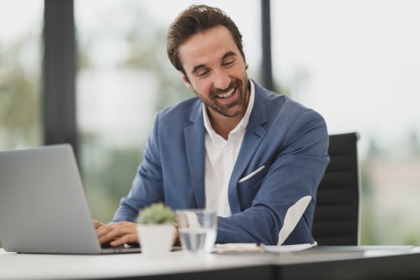 Un hombre de negocios sonriendo mientras trabaja frente a su computadora portátil en una oficina moderna y luminosa.