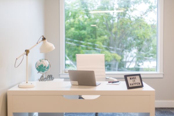 Un escritorio de oficina organizado y moderno con una computadora portátil, una lámpara y un letrero inspirador, ubicado frente a una ventana con vista exterior, representando un ambiente adecuado para el trabajo remoto en una PYME.